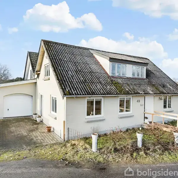 Et hvidt hus med skråt tag og flere vinduer står stille. Ved siden af er en garage. Husnummeret 49 ses på facaden. Omkring er der græs og tæer.