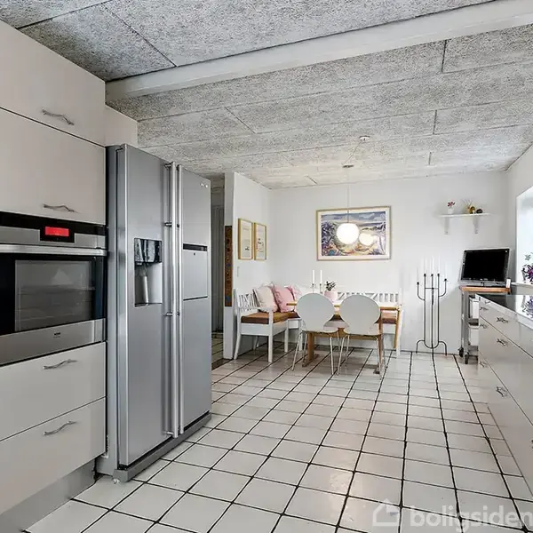 Køkken med stål-refrigerator og indbygget ovn står i forgrunden. En spiseplads med hvide stole og træbord ses bagved, omgivet af køkkenskabe og dekorationer. Ingen tekst.