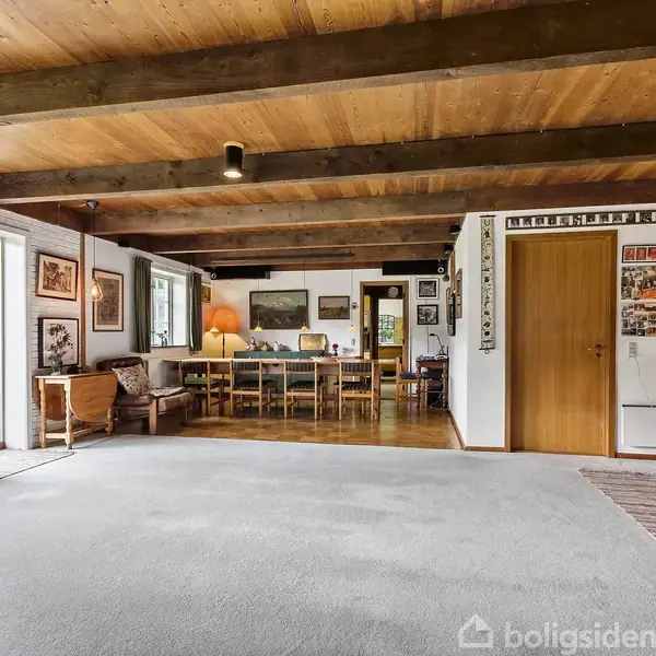 En stue med åbne døre fører ud til en have. I midten er der et spisebord med stole. Rummet har tæppe, træloft, bjælker og mange indrammede billeder på væggene.