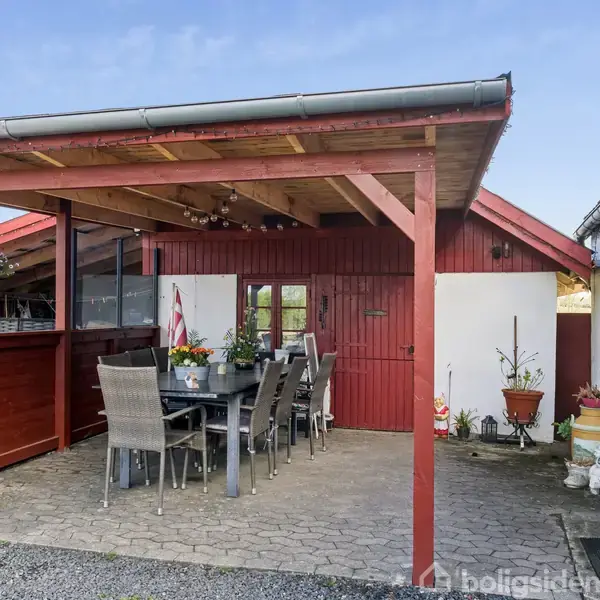 En overdækket terrasse med et spisebord og stole. Terrassen er lavet af træ og fliser i gulvet. Den er omgivet af en rød bygning og blomsterpotter.