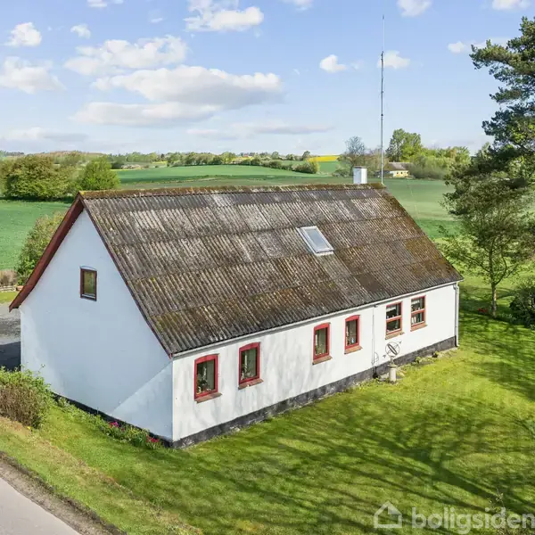 Hvidt hus med røde vinduesrammer står på en grøn græsplæne. Vej løber langs huset; marker og træer er i baggrunden under en delvist skyet himmel.