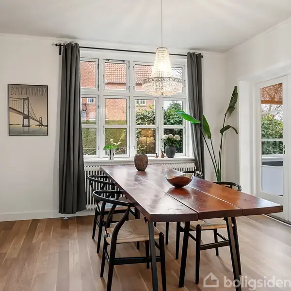 Et træbord med stole står midt i et lyst, velindrettet spisestue med parketgulv. Baggrunden viser store vinduer med grå gardiner og planter. En lysende loftlampe hænger over bordet.