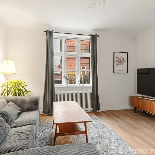 Sofaen står ved siden af en gulvlampe og et sofabord i et lyst stuehjørne med trægulv. En stor fladskærms-TV er placeret på et mediemøbel ved væggen.