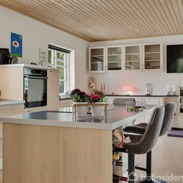 Et moderne køkken har lyse træskabe og en stor ø midt i rummet. Der er barstole ved øen, og overfladerne er dekoreret med planter og køkkenudstyr. I baggrunden ses et stort, fladt fjernsyn på væggen.