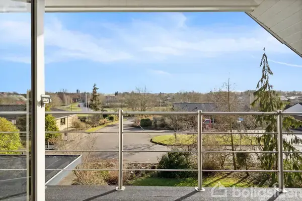 Altan med glasværn kigger ud over bebyggelse og landskab. Det er en solrig dag med en forårsagtig himmel, billedet viser veje, træer og hustage i baggrunden.