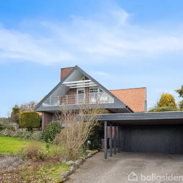 En muret villa med skråt tag og balkon står stille ved siden af en carport. Haven er velplejet med grønne buske og træer, og himlen er klar og blå.