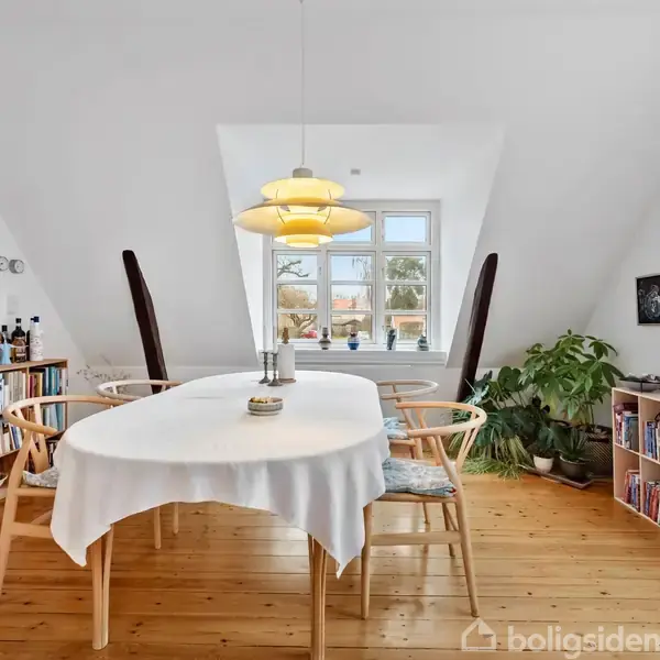 Langbord dækket med hvid dug står i lyst rum; omgivet af stole, reol med bøger, malerier, planter, og et vindue med udsigt til træer. Loftlamper hænger over bordet.