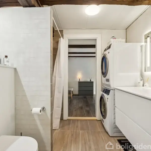 Toilet ved væg, under hylde med toiletartikler; vaskemaskine og tørretumbler stabel i hjørne; dobbeltskab og spejl over vask; åbning til tilstødende rum.