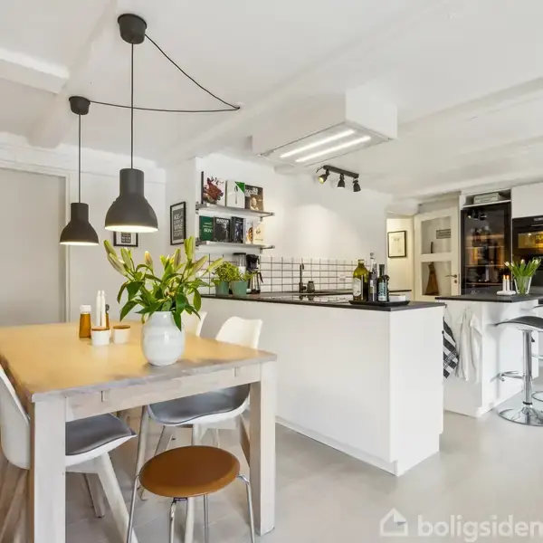 Et lyst køkken-alrum møbleret med en træbord omringet af stole står ved siden af et moderne køkkenområde med sort/hvid indretning og høje barstole. Blomster udsmykker bordet.
