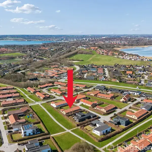Et boligområde med rødt tagede huse, omgivet af grønne områder og veje. En rød pil peger på et specifikt hus, mens en sø kan ses i baggrunden under en blå himmel.
