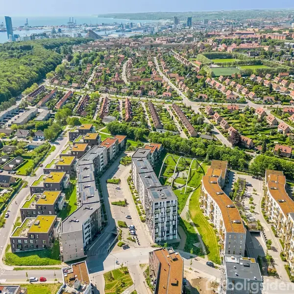 Moderne bygninger med grønne tage danner et boligkompleks. Bygningerne er placeret blandt snoede veje omgivet af skov til venstre og en byudsigten mod havnen i baggrunden.