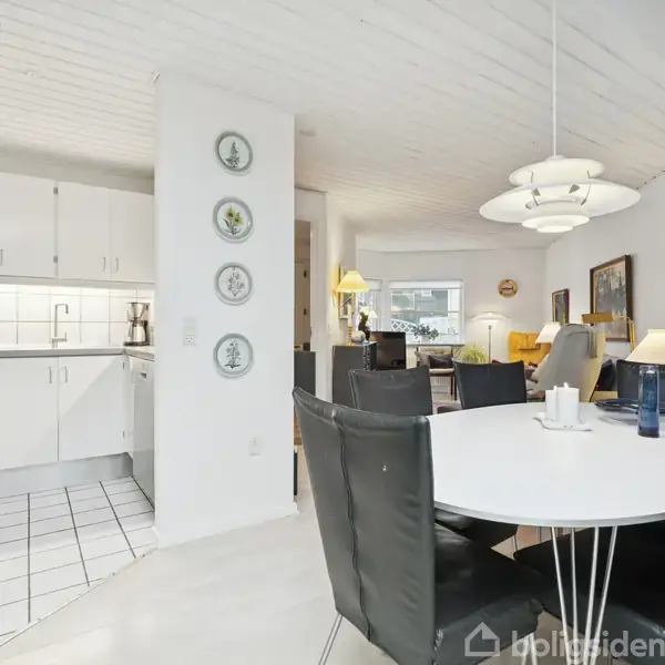 Et rundt spisebord med sorte stole står i et lyst, moderne køkken-alrum. Køkkenet til venstre har hvide skabe. Baggrunden viser en stue med sofaer, lamper og malerier.