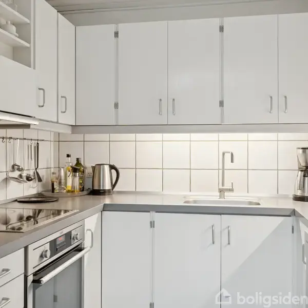 Et hvidt, moderne køkken med skabslåger rundt om. En vask med armatur og en kaffemaskine står på bordet. Fliser dækker væggen over bordpladen. Ovn og kogeplade er indbygget.