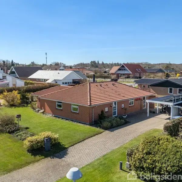 Et rødt murstenshus står på en velholdt græsplæne med fliser i indkørslen. Huset er omgivet af andre huse og plantet buskads under en klar blå himmel.