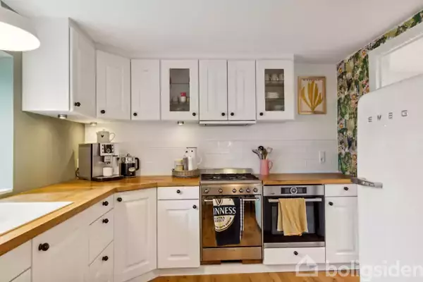 Et lyst køkken med hvide skabe og en træbordplade. Der står en kaffemaskine og brødrister på bordet. En komfurhætte over ovnen med en "Guinness"-håndklæde hængende foran.