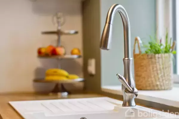 Vandhane i rustfrit stål står ubenyttet ved en køkkenvask. I baggrunden ses en frugtskål med bananer og æbler samt en plante i en fletkurv ved et vindue.