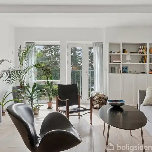 Stue med moderne møbler; sort lænestol og rundt sofabord. Store vinduer lukker naturligt lys ind, omgivet af planter. Hvid bogreol fyldt med bøger og dekorationer.