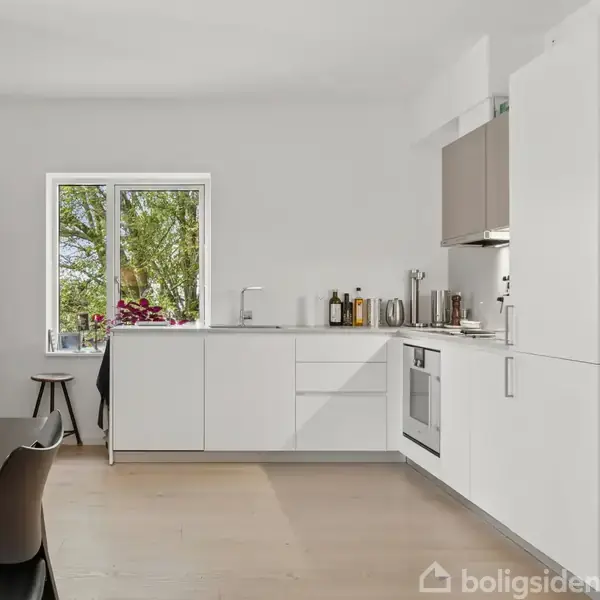 Et lyst køkken med hvide skabe, komfur og køkkenredskaber langs væggen, handling foregår ved et vindue med udsigt til grønne træer, og en sort stol står ved det hvide bord.