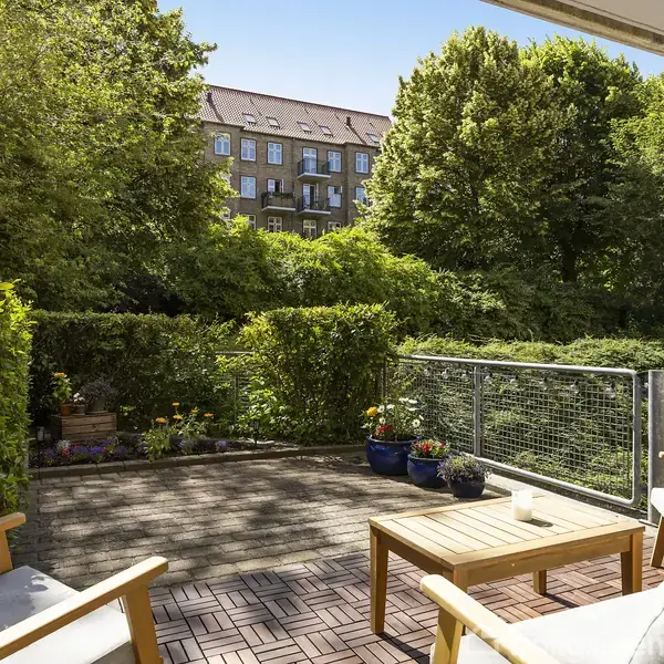 Terrassemøbler med to stole og et bord står på en flisebelagt terrasse. Omgivet af grønne buske og træer med et bygningskompleks i baggrunden.