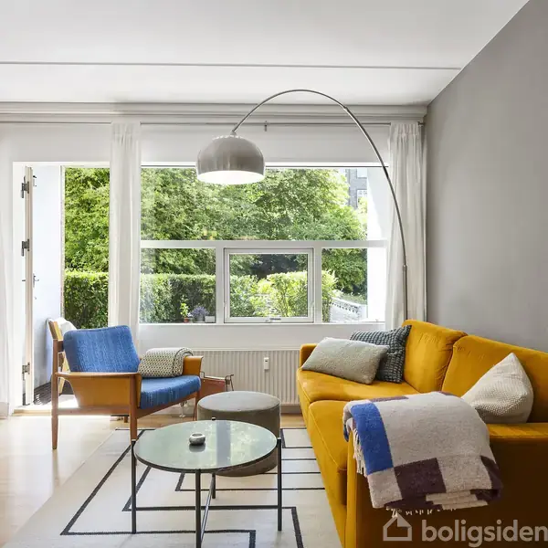 En gul sofa står i en moderne stue. Den er ved siden af et stort vindue med udsigt til grønne træer. Rummet har også en blå lænestol, et sofabord og hylder med bøger.