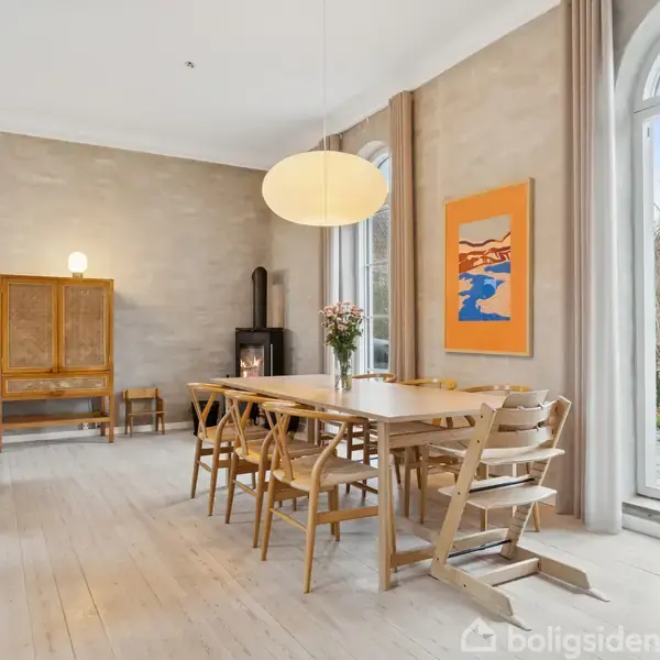 Et langt spisebord med træstole står i en lys stue. En stor lampe hænger over bordet. Der er vinduer mod en have. En reol og brændeovn er i baggrunden.