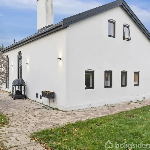 Hvid bygning med sort tag har en muret gangsti omgivet af græsplæne. Der er en udendørs pejs ved væggen. Træer står til venstre under en blå himmel.