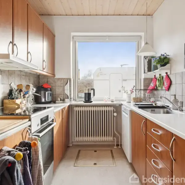 Et lyst køkken med træskabslåger og hvide bordplader. Et vindue over en radiator lader naturligt lys ind. Både venstre og højre side har indbyggede skabe.