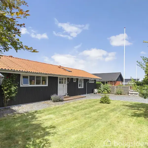 Et sort træhus med rødt tegltag står i en velplejet have med grøn græsplæne og omgivende træer. En flagstang er placeret i baggrunden under en klar blå himmel.