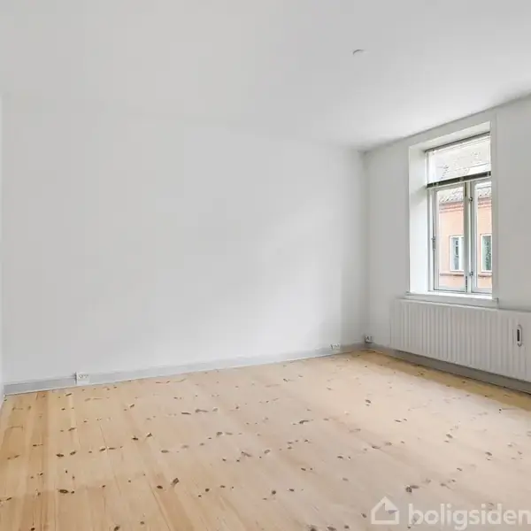 Et tomt værelse med lyse træplanker, hvide vægge, to vinduer med udsigt til bygninger udenfor, og en radiator under vinduerne. En åben dør fører ind til rummet.