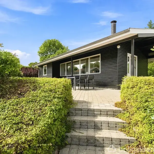 Sort træhus står stille i en frodig have, mens en trappe i forgrunden fører op til en terrasse med havemøbler under en blå himmel.