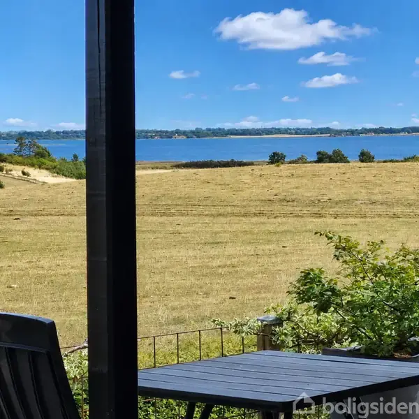 Et bord og stole står på en terrasse med udsigt over en gylden eng, som strækker sig mod en rolig blå fjord og en blå himmel med lette skyer.
