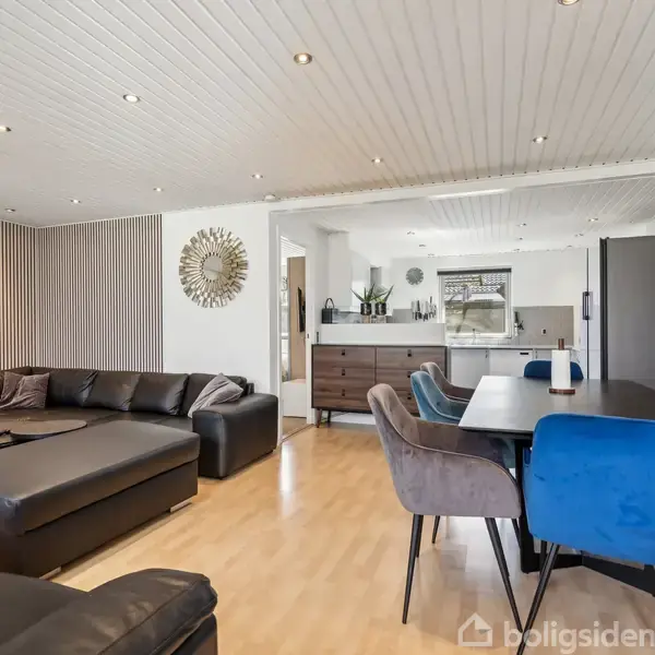 Lædersofa i hjørne; sofabord ved siden. Spisebord med stole foran, midt i opholdsrum. Køkken i baggrunden. Dekorativt spejl og kunstværk hænger på væggen.