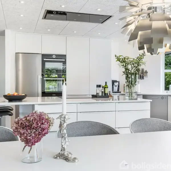 En moderne køkkenalrum med hvide skabe og en stor køkkenø. En dekorativ lysekrone hænger over bordet med blomster og en lysestage. Vinduer giver udsigt til grønne omgivelser.