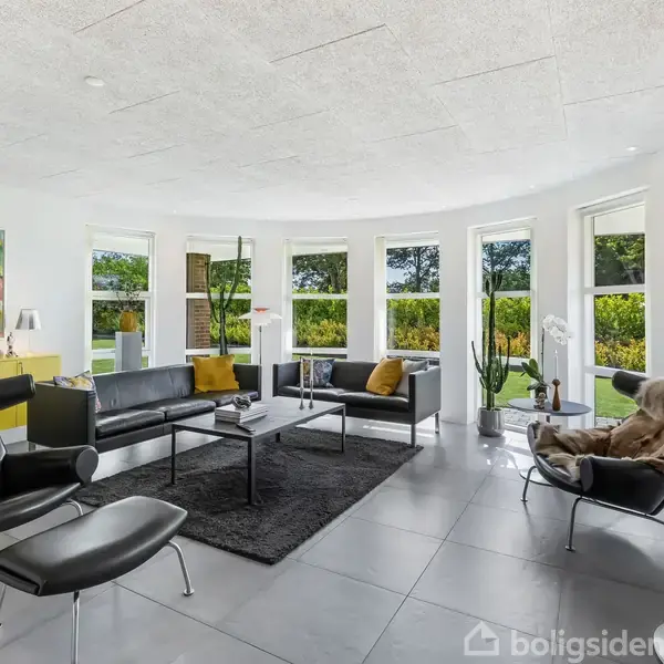 En moderne stue med sorte lædermøbler står foran store, buede vinduer. Et farverigt maleri hænger på væggen over en gul skænk. Grønne planter pynter rummet.