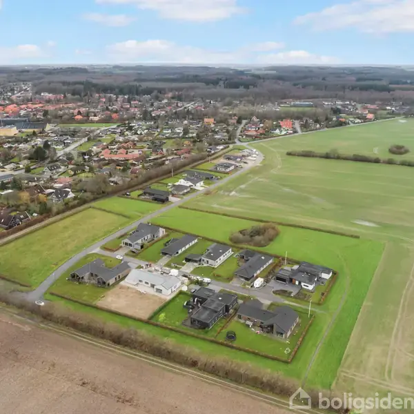 Et lille boligområde med sorte tage står stille, omgivet af store grønne marker på en solrig dag. En større by med flere huse og veje ses i baggrunden.