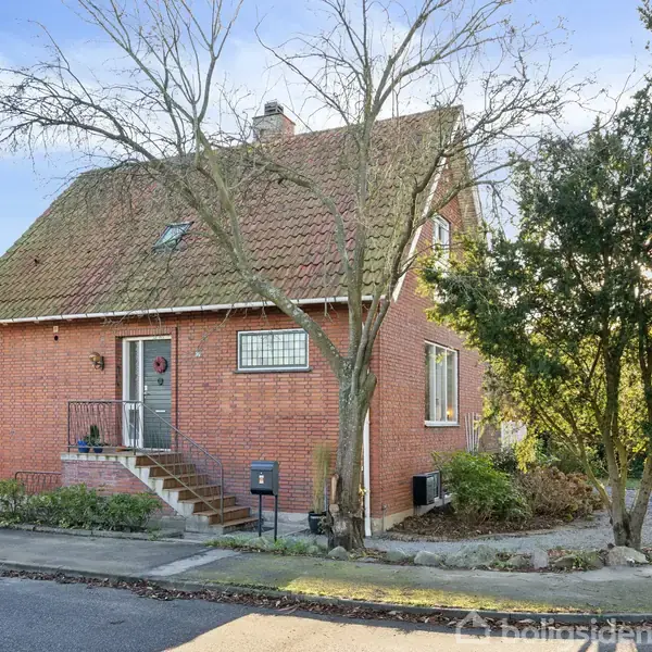 Rødt murstenshus står stille langs en gade; hævede trapper fører til hoveddøren. Omgivet af træer og buske under en blå himmel, skaber et fredeligt boligkvarter.