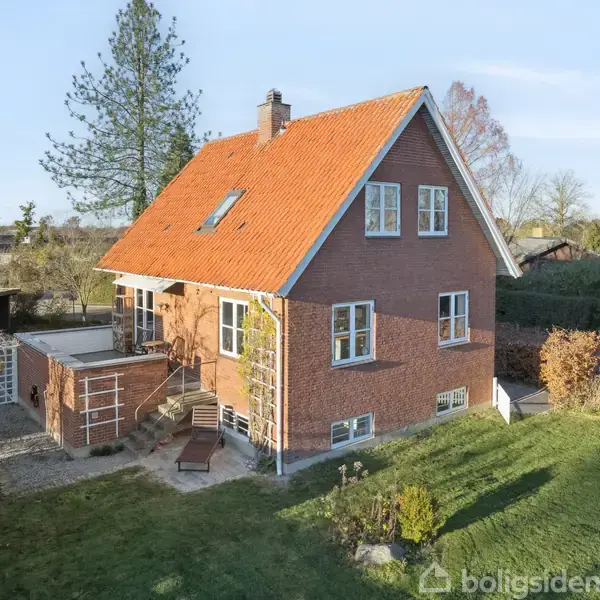 Rødt murstenshus med orange tegltag, omgivet af en grøn have. Huset har flere vinduer og en terrasse med liggestol. Omgivelserne er åbne med træer og buske.