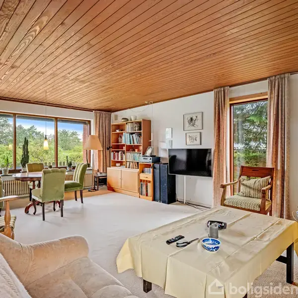 En stue med en beige sofa og bord i forgrunden, fjernsyn på væggen, og spisebord med grønne stole nær vindue. Hylder med bøger og dekorationer langs væggen.