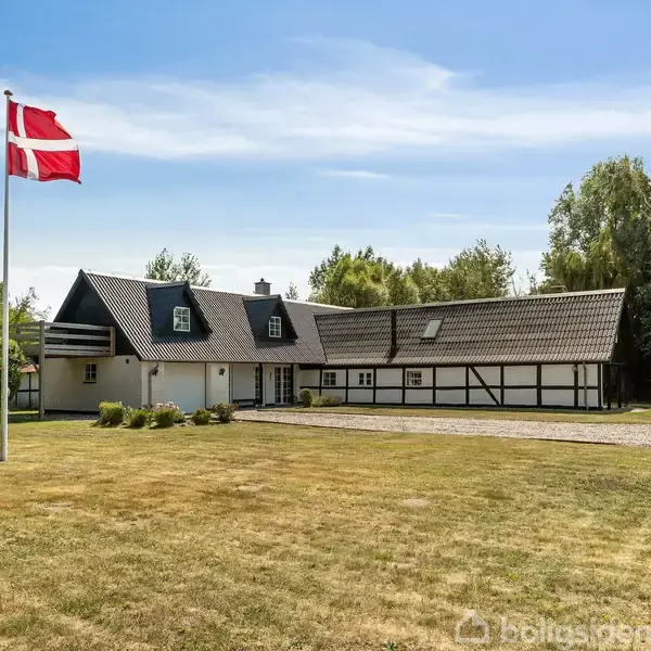 Et hvidt bindingsværkshus med sort tag står i en stor have. Det danske flag vajer i forgrunden, omgivet af græsplæne og træer i baggrunden.