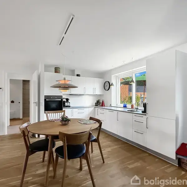 Et moderne køkken med runde spisebord og fire stole står i et lyst rum. Væggen har et stort vindue, hvidt skab og indbygget ovn, mens en lænestol ses til højre.