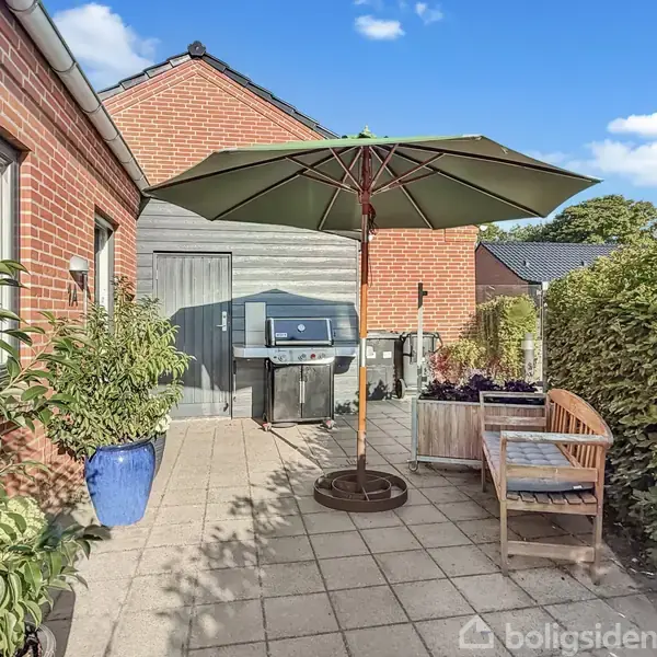 Parasol står åben over en terrasse med flisebelægning. Ved siden af er en bænk og en grill. Omgivet af murstensvæg, planter i krukker og grøn hæk.