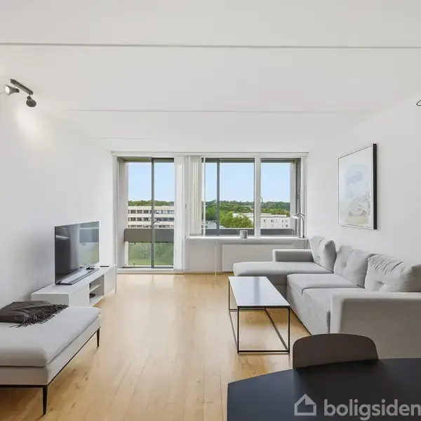 Stue med hvid sofa i hjørnevinkel, fladskærms-TV på lavt bord til venstre, stor vinduesfront med udsigt over grønne områder; lyst træ på gulvet, minimalistisk kunst på væggene.