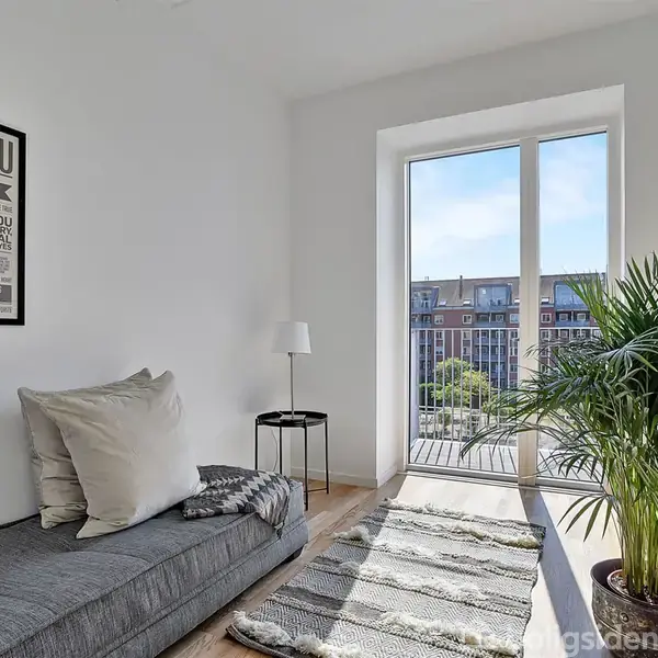 Sofa med puder står i en lys stue, tæt på en glasdør mod en balkon. En plakat med teksten "I LOVE YOU" hænger på væggen. En plante og lampe er ved siden.