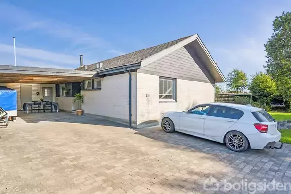 Et hvidt hus med skråtag har en parkeret hvid bil foran indkørslen. Til venstre er en overdækket terrasse med bord og stole. Grønne træer omkranser baggrunden.