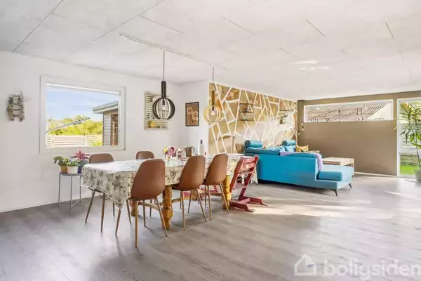 Spisebord med blomsterpynt står ved siden af en blå sofa i en moderne stue med trægulv, geometriske vægdetaljer og store vinduer, der slipper naturligt lys ind.