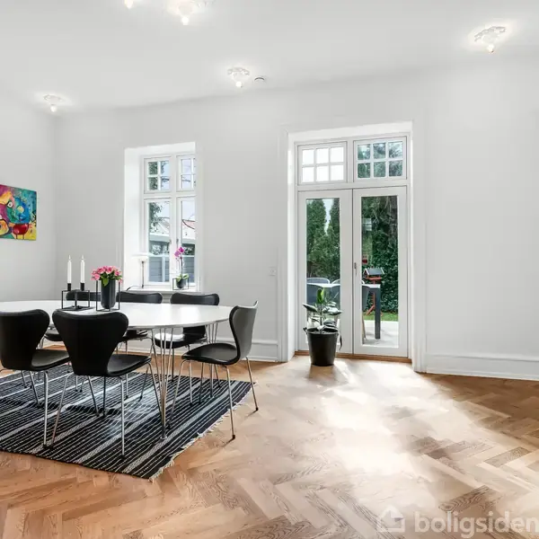 Spisebord med sorte stole står på et tæppe, omgivet af hvide vægge og store vinduer med udsyn til en have. Et maleri hænger på væggen, og en plante står ved døren.