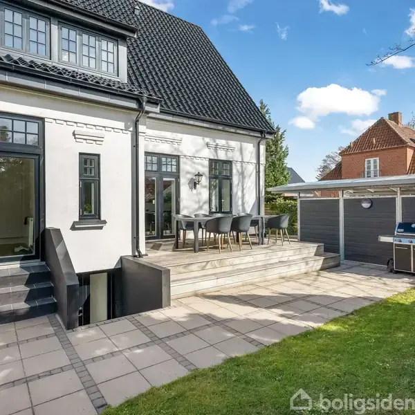 Et hus med sort tag har en stor terrasse med havemøbler. Trin fører ned til en flisebelagt gårdhave. En carport med grill og redskaber står til højre.