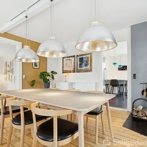 Et lyst spisebord med seks stole står under tre store lamper. I baggrunden er en moderne brændeovn med træstabler ved siden af. Rummet har hvide vægge og trægulv.