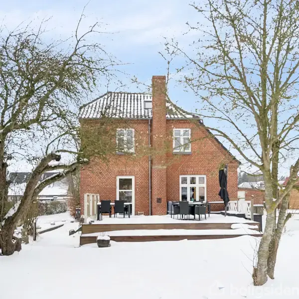 Rødt murstenshus står stille i snedækket have med træer, terrassemøbler og høj blå himmel.