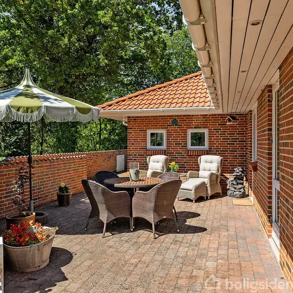 Terrasse med kurvemøbler omkring et rundt bord og en parasol. Omkring står blomsterkrukker. Omgivet af rød murstensvæg og grønne træer, skaber et hyggeligt udeområde.